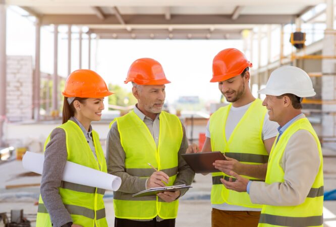 Meeting. A smiling and positive group of architectures standing together and specking about construction project with construction works on a background