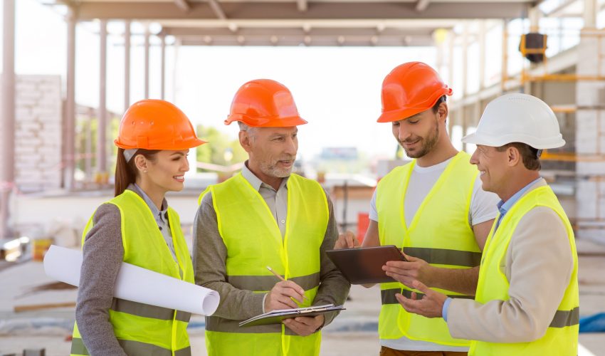 Meeting. A smiling and positive group of architectures standing together and specking about construction project with construction works on a background